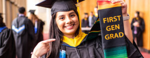 Graduate smiling while holding a First Gen Grad stole during commencement.